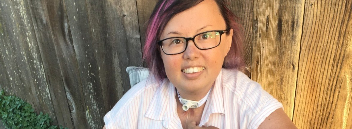 Image Description: Stacy Milburn, Korean queer disabled activist sitting in her chair with black rimmed glasses, pink hair, and a pink striped shirt. She is smiling at the camera and looking joyful.