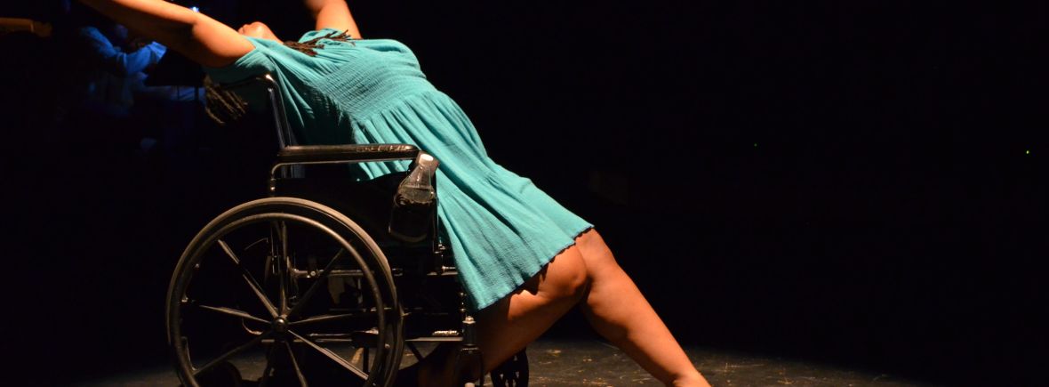 India Harville, African-American disabled dancer using a wheelchair is leaning back on her chair with her arms reaching behind her. She is wearing a teal dress and new shoes. Her head is not visible and is behind her arms.